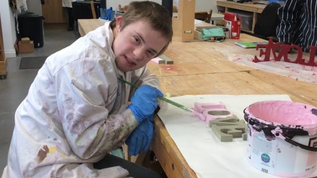 Student smiling whilst painting a wooden ornament saying love