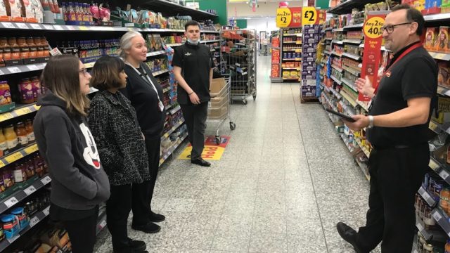 Students and staff listening to Morrisons team leader deliver training induction in the supermarket aisle