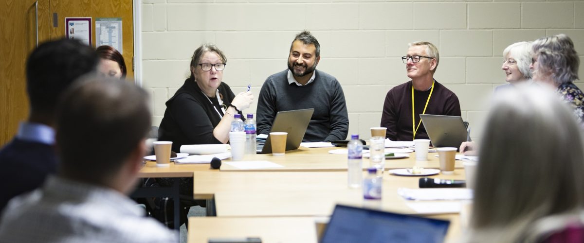 9 people around a meeting table mid discussion