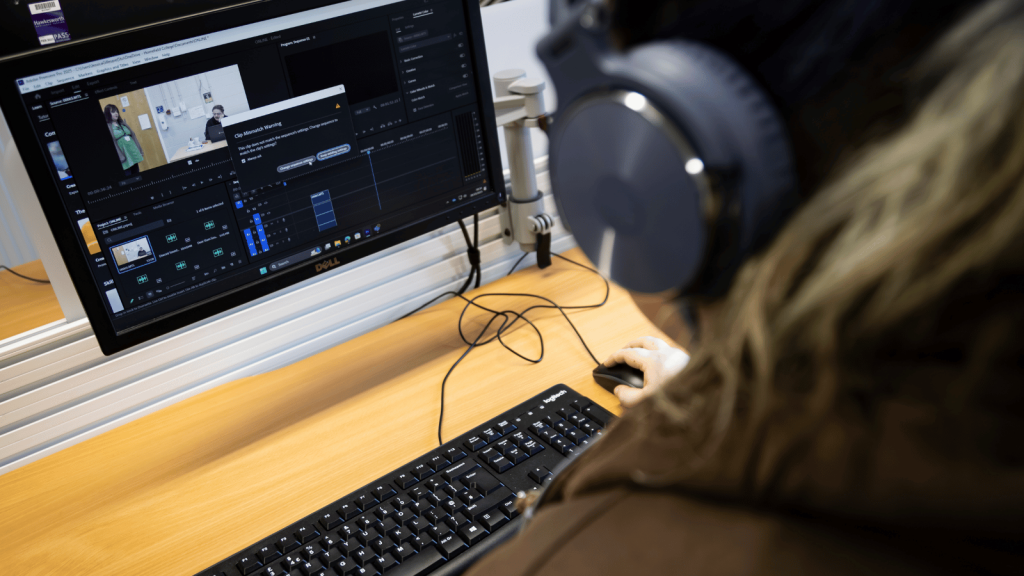 Image of student Jess wearing headphones and working on editing a video on her monitor using Adobe software.