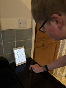 Student Corwin looking at his iPad screen, which shows his washing up task list: "Put the plug in the sink. Add a few drops of washing up liquid. Fill the sink with warm water. Put the pot in the water. Scrub with the sponge."