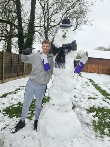 Student Jacob posing cheerfully next to a tall snowman decorated with several household items: a hat, a scarf, gloves, and various vegetables used to create the face.