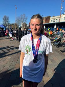 A photo of Emily smiling at the camera, wearing a Homefield t-shirt and her Leicester 10K medal.