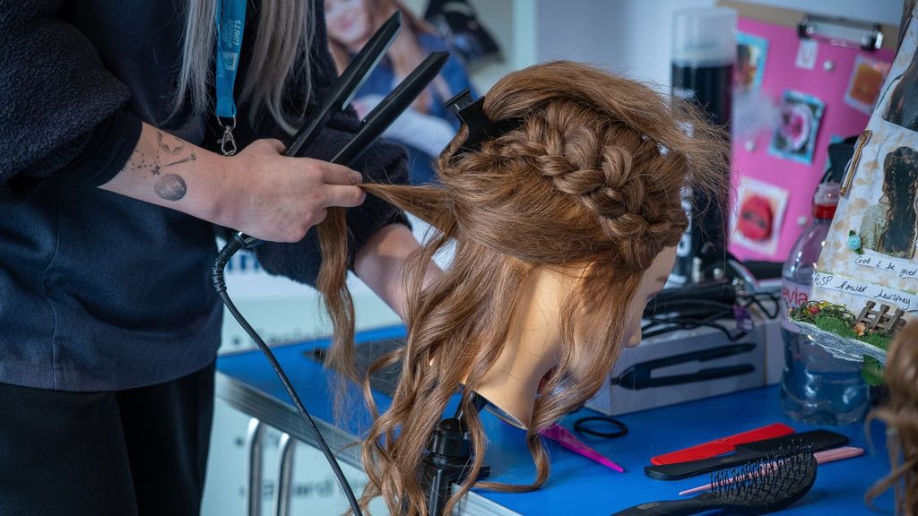A photo of a student's hands as they hold a hair straightener and style a mannequin head's hair